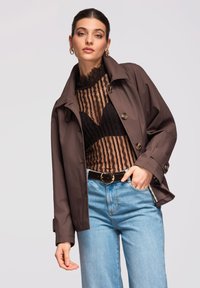 Femme portant un manteau marron boutonné par-dessus un haut en dentelle noire transparente, un jean bleu avec une ceinture noire et des boucles d'oreilles dorées en forme d'anneau, posant devant un fond uni.