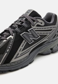 Juodas ir pilkas sportinis batelis su tinkline viršutine dalimi, paminkštintu apvadu ir tekstūruota padais. Yra atspindinčio logotipo ir išskirtinio kampinio dizaino.