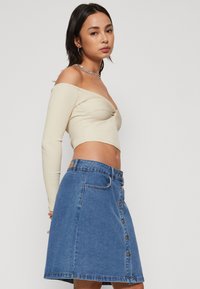 Krém színű, bordázott, váll nélküli crop top hosszú ujjal, párosítva egy kék farmer A-vonalú szoknyával, elülső gombokkal és klasszikus zsebekkel.