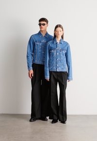 Két modell visel kék farmer dzsekit, amelyen négy első zseb található, fekete bő szárú nadrággal párosítva. A környezet minimalista.