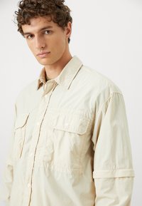 Camicia beige chiaro realizzata in un tessuto leggero, con chiusura a bottoni sul davanti, due tasche sul petto e maniche arrotolate. Design casual e texture interessante.