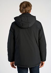 Musta pehmeä parkatakki hupulla, jossa on sileä pinta ja hieman pidempi leikkaus, näkyvät ompeleet ja joustavat hihansuut.