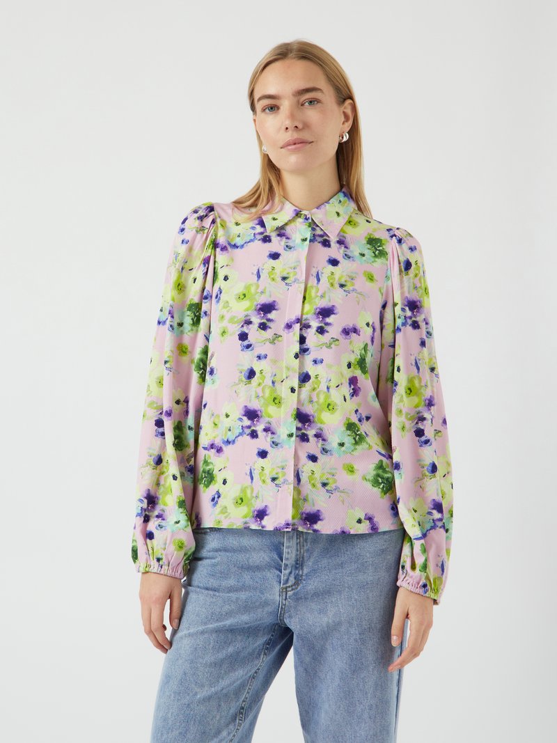 Blusa floral em rosa claro com padrões verdes e roxos, apresentando um colarinho, mangas bufantes e punhos elásticos. Combinada com jeans de ganga.
