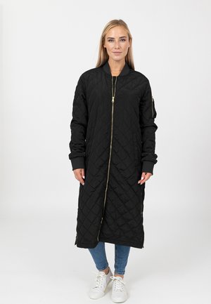 Zwarte gewatteerde lange jas met een slim fit en gouden rits. Voorzien van ribgebreide manchetten en zakken. Gedragen met blauwe spijkerbroek en witte schoenen.