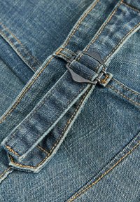 Džinsinio audinio artimas vaizdas, kuriame matyti metalinė sagtis, užseganti reguliuojamą dirželį su oranžiniais siūlų siūlėmis.