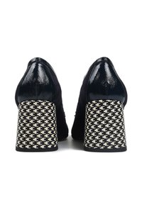 Para czarnych, zamszowych i lakierowanych butów na słupku, z obcasami w czarno-białą kratę houndstooth, widziana od tyłu.