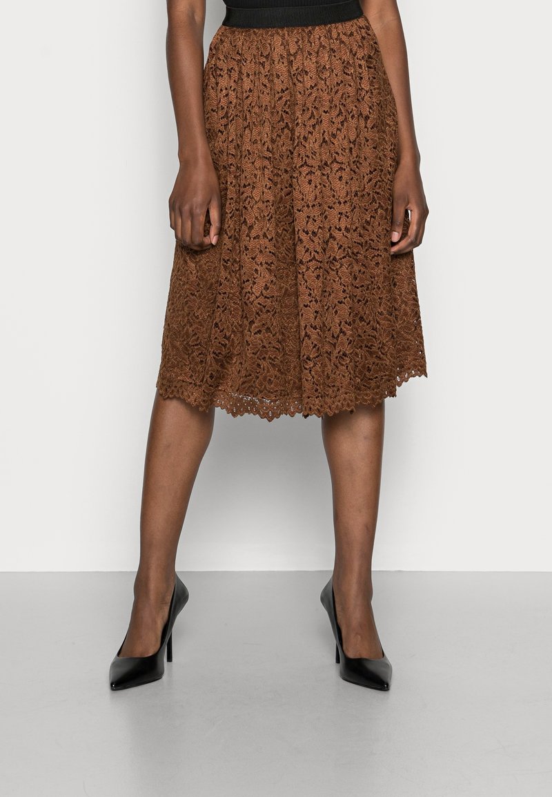 Bruine midi-rok van kant met bloemenpatroon, gegolfde zoom en een aansluitende zwarte tailleband, gecombineerd met zwarte, hoge hakken.