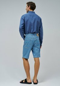 Bermudas de mezclilla azul claro con un corte clásico, que presentan un diseño de bolsillo trasero y una etiqueta marrón. Combinadas con una camisa azul de botones.