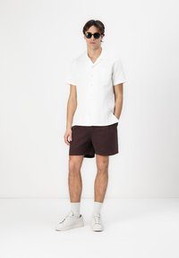 Camisa blanca de botones de manga corta con un patrón texturizado, combinada con pantalones cortos marrón oscuro y zapatillas blancas. Las gafas de sol completan el look.