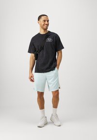 Camiseta negra de gran tamaño con logo blanco, pantalones cortos de malla azul claro, calcetas blancas y zapatillas blancas. El modelo sostiene una bolsa de hombro negra.