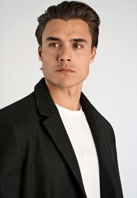 Abrigo de lana negro con solapas muesca, acabado texturizado, llevado sobre una camiseta de cuello redondo blanca. El modelo tiene el cabello corto y peinado, y un fondo neutro.