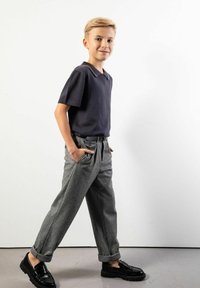 Blond chłopiec idzie z rękami w kieszeniach szarych spodni, nosząc czarne mokasyny i ciemny krótki sweter z krótkim rękawem na białym tle.