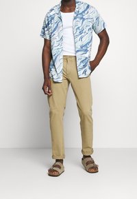 Camicetta a maniche corte, di colore azzurro chiaro con un motivo marmorizzato a spirale, abbinata a pantaloni beige e sandali marroni con cinghie regolabili.