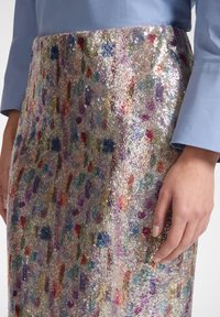 Blå skjorte med lange ærmer, stoppet ind i et multifarvet palietnederdel, med en hånd hvilende blidt på siden af nederdelen.