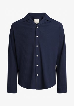 Camicia a maniche lunghe blu navy in tessuto a coste, con colletto aperto, bottoni frontali e un'etichetta sul lato sinistro in basso.