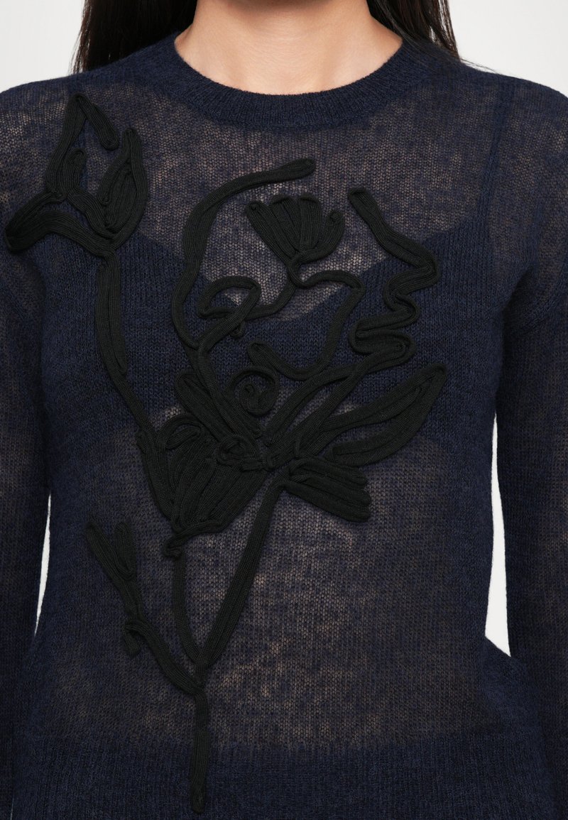 Pullover trasparente blu navy con ricami floreali astratti neri a texture sulla parte anteriore, indossato da una persona con lunghi capelli scuri.