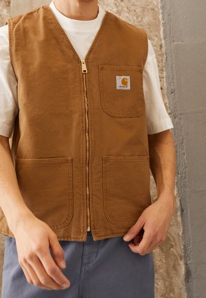 Homme portant un gilet sans manches marron zippé avec poches avant par-dessus un t-shirt blanc, debout contre un mur beige et gris texturé.