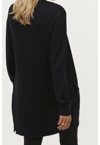 Kvinna som bär en lång svart cardigan med framfickor, svarta byxor och en guldring, sedd från ryggen mot en neutral bakgrund.