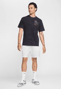 Black cotton t-shirt with a textured finish and a patterned pocket detail, paired with white athletic shorts and black-and-white sneakers.