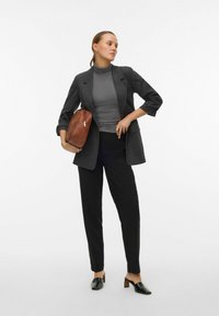 Grå ternet blazer over en grå turtleneck, sorte bukser og sorte hælede mule sko. Brun skuldertaske med lynlåsdetalje. Ren baggrund.