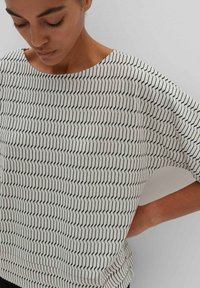 White short-sleeve top with geometric black zigzag pattern, made of lightweight fabric with a loose fit and rounded neckline.