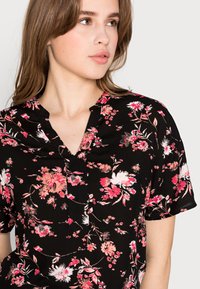 Blusa nera a fiori con fiori rosa e bianchi, maniche corte, scollatura a V e una texture sottile. Presenta un design a bottoni sulla scollatura.