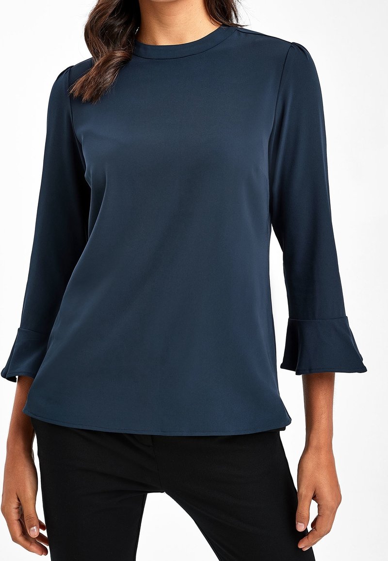 Námořnický modrý top s tříčtvrtečními rukávy, se zvonovými manžetami a hladkým povrchem. Kulatý výstřih a volný střih. Viditelné černé kalhoty.