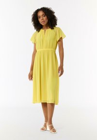 Gelbes knielanges Kleid mit gerafftem Bund und Rüschensäumen; leichtes Material; Schlüssel-Ausschnitt; getragen mit weißen Sandalen mit Riemchen.