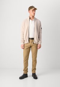 Lys beige ribbet cardigan over en hvit polo skjorte, parret med lysebrune chinos og svarte ankelstøvler. En mørkegrønn caps fullfører looket.