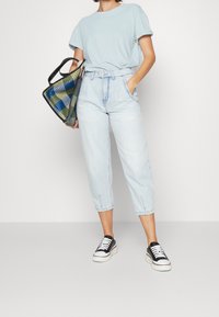 Maglietta azzurra, jeans in denim chiaro a vita alta e scarpe da ginnastica bianco e nero. Il modello tiene una borsa a tracolla nera con un motivo.