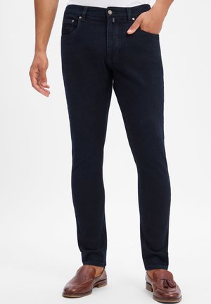 Uomo che indossa jeans slim fit blu scuro e mocassini in pelle marrone, in piedi con una mano in tasca.