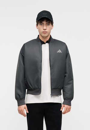 Jeune homme portant une casquette noire adidas, une veste bomber gris foncé avec logo, une chemise blanche et un pantalon noir, debout devant un fond clair uni.