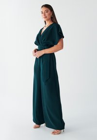 Zaļš, plašu kāju jumpsuits ar dziļu V-veida izgriezumu, īsām plandošām piedurknēm un sasienamu vidukli. Modelis valkā zelta krāsas sandales ar siksniņām.