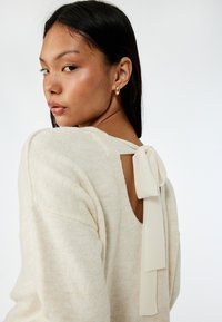 Crème sweater met een brede scoop rug en een gestrikte strikdetail bij de hals. Zachte, gestructureerde stof met een losse pasvorm. Ronde mouwen.