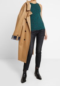 Tyrkysový tílko s černým leopardím vzorem, béžový trenčkot, tmavé úzké džíny a černé kotníkové boty. Trenčkot má kostkované manžety.