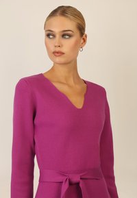 Fuchsia Wollsweater mit V-Ausschnitt, langen Ärmeln und einem gebundenen Taillendetail, das eine glatte Textur und eine taillierte Silhouette aufweist.