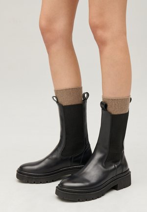 Botas de tobillo negras de cuero con paneles laterales elásticos texturizados y pestañas, que cuentan con una suela gruesa y calcetines de punto marrón acanalados.