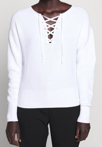 Maglione bianco lavorato a maglia con maniche lunghe, orlo a costine e dettagli con lacci sul collo. Texture morbida e vestibilità comoda.