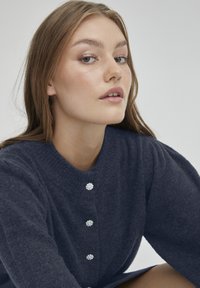 Mörkblå tröja med ribbad halsringning och fyra dekorativa rhinstenknappar framtill. Mjuk textur och något puffade ärmar.