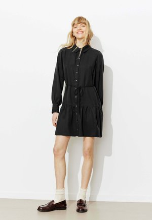 Femme aux longs cheveux blonds sourit, portant une robe noire boutonnée à manches longues, des chaussettes blanches aux chevilles et des mocassins marron foncé, debout contre un mur blanc.