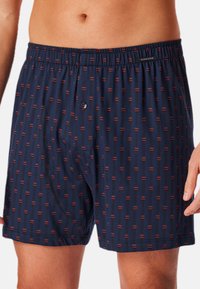 Marineblauwe boxers met een herhalend patroon van rode en zwarte verticale lijnen. Heeft een elastische tailleband en een knoopsluiting aan de voorkant.