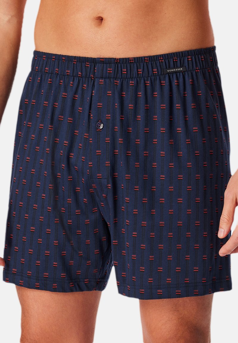 Marineblauwe boxers met een herhalend patroon van rode en zwarte verticale lijnen. Heeft een elastische tailleband en een knoopsluiting aan de voorkant.