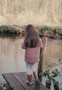 Enfant portant une veste polaire rose et des leggings blancs se tenant sur un quai en bois près de l'eau, entouré de verdure.