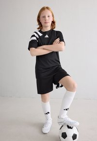 Μαύρη ποδοσφαιρική στολή με λευκές ρίγες, λευκά κάλτσες και λευκά παπούτσια. Σταθεί σε μια μαύρη και λευκή μπάλα ποδοσφαίρου, με τα χέρια σταυρωμένα αυτοπεποίθητα.