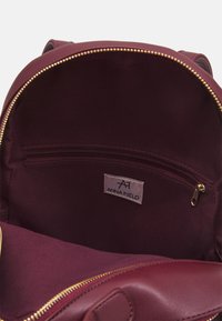 Sac à dos bordeaux ouvert avec une fermeture éclair dorée, révélant une poche intérieure zippée et une étiquette en tissu indiquant "Anna Field".