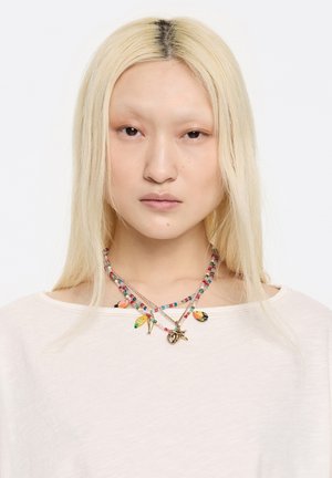 Collane di perline colorate con charm a forma di pesci e uccelli; perline multicolori su catene multiple, esposte su una maglietta leggera e ampia.