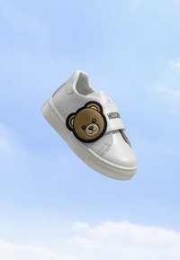 Бели детски маратонки с декорация на плюшено мече и лого на Moschino на каишката на фона на синьо небе.