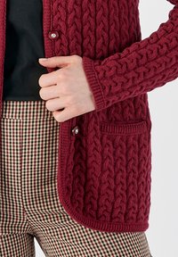 Gilet en tricot bordeaux avec un motif en torsades, doté d'une fermeture à boutons et d'une poche à l'avant, associé à un pantalon à carreaux dans des tons neutres.