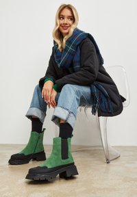 Groene suède enkellaarzen met zwarte rubberen zolen, voorzien van zijritsen en een chunky ontwerp. Model draagt opgerolde jeans en een blauwe geruite sjaal.