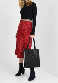 Sac tote noir avec un accent métallique circulaire, fabriqué en matériel lisse. Le sac présente des poignées doubles et une forme rectangulaire structurée.